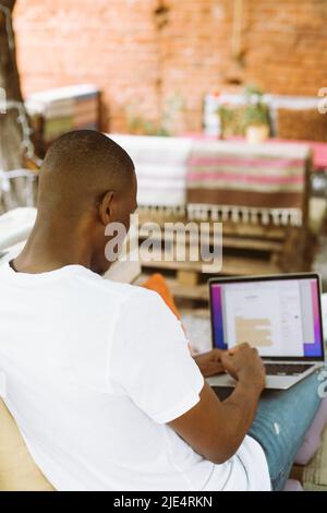 Rückansicht konzentriert afroamerikanischen Mann mit Laptop sitzen auf Bank mit Beinen im Café im Freien. Bearbeiten von Verträgen Stockfoto