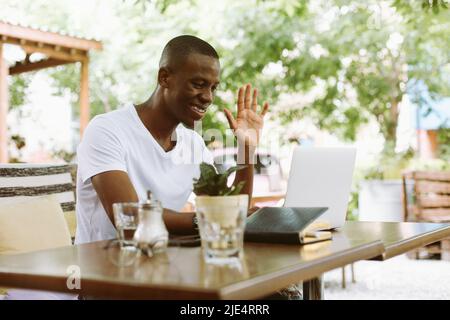 Lächelnder und fröhlicher multirassischer Mann mit Laptop, der den Arm hebt, jemanden begrüßt und auf den Monitor schaut. E-Learning-Webinar-Meeting Stockfoto