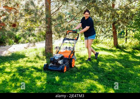 Müde braunhaarige Frau in legerer Kleidung mit Benzin-Rasenmäher oder Grasschneider Pflege ihren Rasen auf dem Land Stockfoto