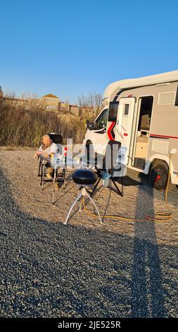 Reisemobil Reisen. Frau trinkt Morgenkaffee vor dem Wohnmobil, im Vordergrund ein Grill Stockfoto