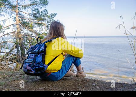 An bewölktem Tag im Frühjahr sitzt eine junge Frau mit Rucksack an der Küste des Waldsees. Zeit für sich selbst. Kontemplation und Ruhe in Einsamkeit Stockfoto