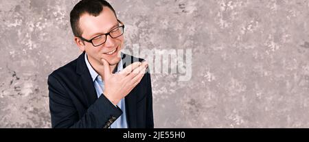 Porträt eines jungen lächelnden modernen Geschäftsmannes in einer Brille in einem Anzug vor dem Hintergrund einer grauen Betonwand. Platz zum Kopieren, Platz für Text Stockfoto