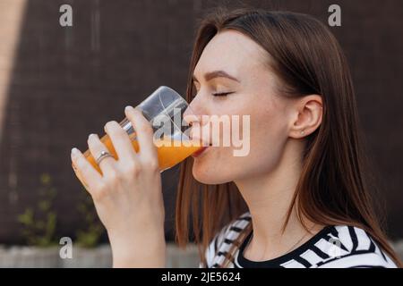 Porträt einer schönen jungen Frau, die draußen auf der Terrasse frischen, kalten Orangensaft trinkt und trinkt, Nahaufnahme von der Seite. Dame mit geschlossenen Augen Stockfoto