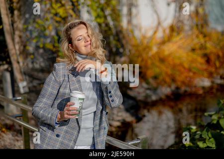 Genießen Sie den Moment. Charmante Blondine mit langen Haaren in hellgrauem Kaschmirpullover und Jacke, passend zu heißem Kaffee in der Tasse, glücklich am sonnigen Herbsttag Stockfoto
