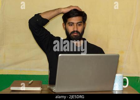 Ein bärtiger indischer Mann in einem schwarzen formellen Hemd, der auf einem Stuhl sitzt und einen schockierten Blick auf den Laptop-Bildschirm gibt. Tagebuch und Kaffeebecher auf ta Stockfoto