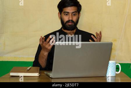 Ein bärtiger indischer Mann in einem schwarzen formellen Hemd, der auf einem Stuhl sitzt und einen überraschten Ausdruck beim Blick auf den Laptop-Bildschirm gibt. Tagebuch und Kaffeebecher an Stockfoto
