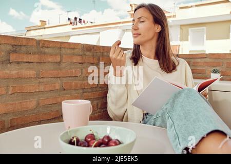 Junge schöne Frau, die sich auf der Terrasse ihres Hauses entspannt und Kirschen isst. Stockfoto