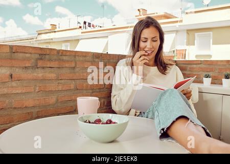 Junge schöne Frau, die sich auf der Terrasse ihres Hauses entspannt und Kirschen isst. Stockfoto