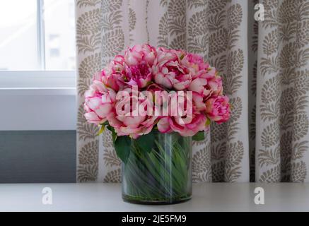 Rosa Blumen in Glasvase mit grünen Stielen zeigt sitzen auf Regal an Fenster mit Muster Stoffvorhang Stockfoto