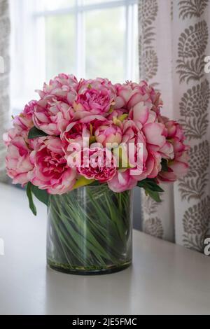 Rosa Blumen in Glasvase mit grünen Stielen zeigt sitzen auf Regal an Fenster mit Muster Stoffvorhang Stockfoto