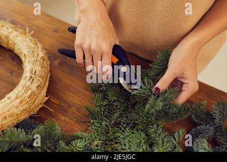 Nahaufnahme einer nicht erkennbaren weiblichen Floristin, die am Holztisch steht und Nadelzweige schneidet, während sie einen Weihnachtskranz anrichtet Stockfoto