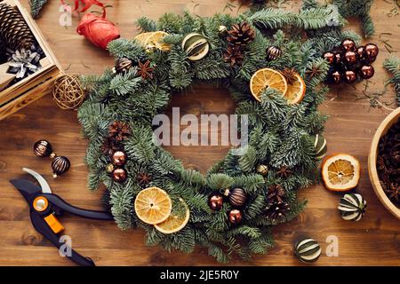 Blick aus dem hohen Winkel auf einen schönen Urlaubskranz mit orangefarbenen Scheiben, Tannenzapfen und kleinen Kugeln, die zwischen Dekorationen und Werkzeugen auf einem Holztisch platziert sind Stockfoto