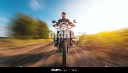 Ein Typ auf einem Motorrad fährt an einem sonnigen Tag mit Geschwindigkeit auf einer unbefestigten Straße. Preisgünstige Motorradbewegungen mit einem Unschärfeeffekt Stockfoto