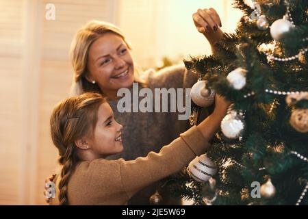 Glückliche schöne Mutter und Tochter schmücken Nadelbaum mit Kugeln während der Vorbereitung nach Hause für Weihnachten Stockfoto