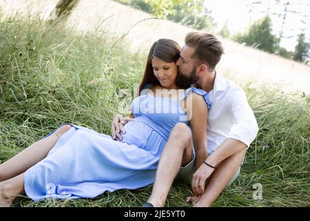 Das verliebte junge Paar sitzt im Sommer auf einer hohen Blumenwiese und kuschelt, die Frau ist schwanger und der Mann hat einen vollen Bart Stockfoto