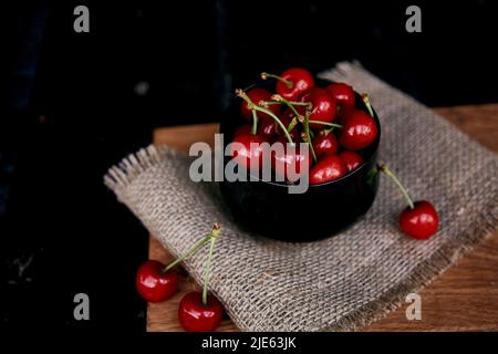 Kirschen in Glasschüssel auf dem Sacking auf schwarzem Hintergrund. Rustikale Bio-Beeren aus eigenem Anbau. Gemütliches Abendessen auf dem Land. Stockfoto