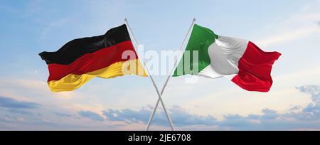 Zwei gekreuzte Flaggen Bundesrepublik Deutschland und Italien winken im Wind bei bewölktem Himmel. Konzept der Beziehung, des Dialogs, des Reisens zwischen zwei Ländern Stockfoto