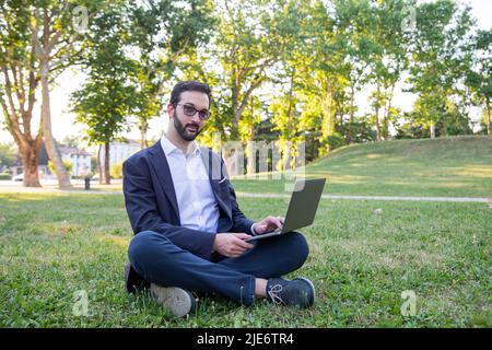 Der junge Unternehmer im Park beantwortet eine E-Mail, während er auf dem Rasen sitzt. Arbeiten Sie überall. Stockfoto