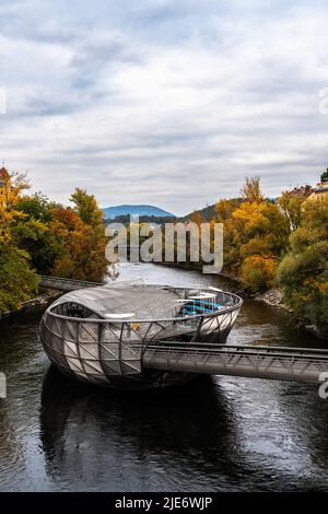 Schöner Blick auf die Murinsel an der Mur mit bunten Bäumen und Bergen im Hintergrund an einem bewölkten Herbsttag, Graz, Österreich Stockfoto