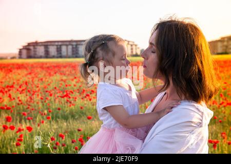 Mama und ihre kleine Tochter in weißen Kleidern posieren in einem Mohnfeld unter blühenden Mohnblumen bei Sonnenuntergang. Rote Mohnblumen und eine schöne Frau mit einem b Stockfoto