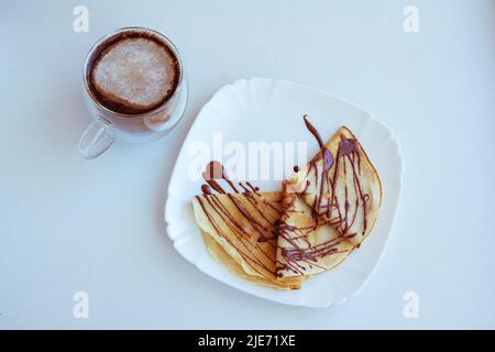 Frischer Kaffee mit Milch in einem Glaskrug und Pfannkuchen mit Schokolade auf einem weißen Teller Stockfoto