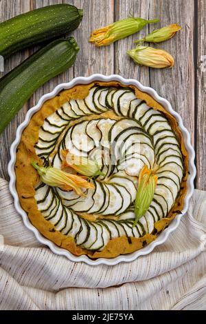 Köstliche hausgemachte Zucchini-Torte mit Quark und Kräutern, umgeben von Blumen, Samen und Zucchini auf einem rauen Holztisch. Draufsicht mit Kopierbereich Stockfoto
