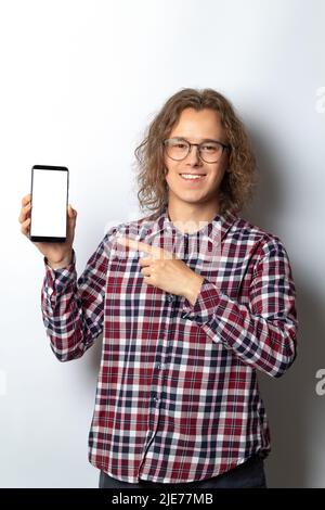 Hübscher junger Mann mit Smartphone und weißem leeren Bildschirm. Der Typ zeigt mit dem Finger auf das leere Handy-Mockup. Stockfoto