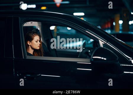 Horizontales Foto von der Seite, nachts, von einer Frau, die in einem schwarzen Auto sitzt und nachdenklich nach vorne schaut Stockfoto