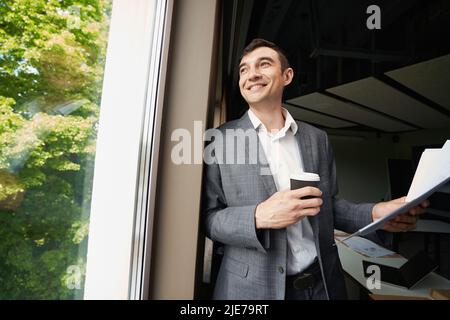 Zufriedener männlicher Büroangestellter mit Kaffee, der ins Fenster schaute Stockfoto