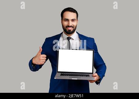 Porträt eines zufriedenen, entzückten bärtigen Mannes, der mit einem Lächeln auf dem Laptop arbeitet und den Daumen nach oben zeigt, die Kamera anschaut und einen Anzug im offiziellen Stil trägt. Innenaufnahme des Studios isoliert auf grauem Hintergrund. Stockfoto