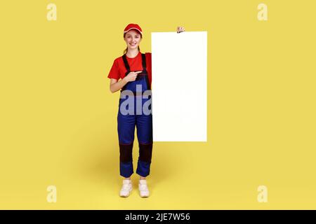 In voller Länge Porträt einer Arbeiterin, die auf ein weißes Papierplakat mit leerem Platz für Werbung oder Werbetexte zeigt. Innenaufnahme des Studios isoliert auf gelbem Hintergrund. Stockfoto