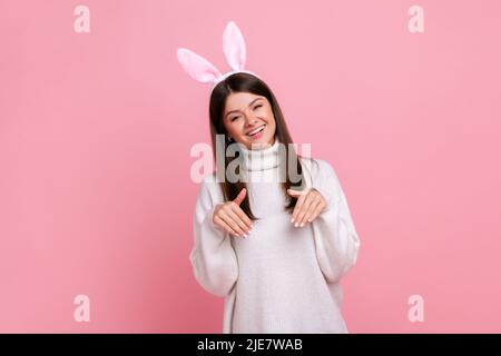 Lustige sorglose junge Erwachsene Brünette Frau mit Hasenohren, schauen verspielt glücklich, kindhaftes Verhalten, tragen weißen lässigen Stil Pullover. Innenaufnahme des Studios isoliert auf rosa Hintergrund. Stockfoto