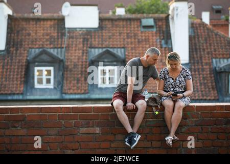 Warschau, Polen. 25.. Juni 2022. Ein Paar schaut auf das Smartphone, während es an den alten Backsteinmauern in der Nähe des Warschauer Barbican sitzt. Tägliches Leben am Sommerabend in der Warschauer Altstadt nach einem heißen Samstag. (Foto von Volha Shukaila/SOPA Images/Sipa USA) Quelle: SIPA USA/Alamy Live News Stockfoto