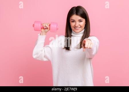 Lächelnde Frau, die Rose mit Hanteln hält und mit dem Finger auf die Kamera zeigt, ruft zum Sport auf und trägt einen weißen Pullover im lässigen Stil. Innenaufnahme des Studios isoliert auf rosa Hintergrund. Stockfoto