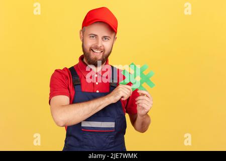 Porträt eines positiv begeisterten Arbeiters, der blaue Overalls trägt, den grünen Hashtag hält und die Kamera mit fröhlicher Ausdruckskraft anschaut. Innenaufnahme des Studios isoliert auf gelbem Hintergrund. Stockfoto