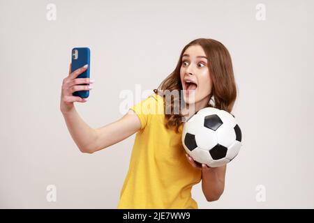 Überglücklich aufgeregt Bloggerin jungen Alters in gelbem Casual T-Shirt mit Fußball in der Hand halten und Live-Stream von Fußballspiel. Innenaufnahme des Studios isoliert auf grauem Hintergrund. Stockfoto