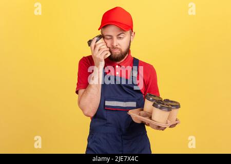 Müde erschöpfte Arbeiter fühlt sich schläferig nach harter Arbeit stehen mit geschlossenen Augen, ein Nickerchen, halten Einweg-Tassen Kaffee, tragen Overalls und Mütze. Innenaufnahme des Studios isoliert auf gelbem Hintergrund Stockfoto