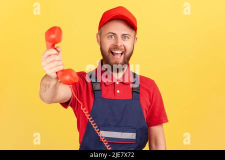 Porträt eines positiven Handyman trägt blaue Uniform halten Hörer, fragen rufen Sie ihn und bestellen Service, Blick auf die Kamera glücklich Ausdruck. Innenaufnahme des Studios isoliert auf gelbem Hintergrund. Stockfoto