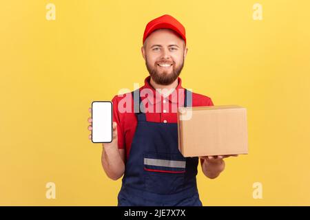 Bärtiger Lieferer hält einen Karton mit bestellten Online-Waren, zeigt Handy mit leerem Bildschirm für Werbung, trägt blaue Overalls. Innenaufnahme des Studios isoliert auf gelbem Hintergrund Stockfoto