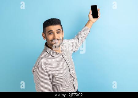 Porträt eines Geschäftsmannes, der Videoanruf unterhielt, Selfie auf dem Mobiltelefon machte, Smartphone mit leerem Modup-Display hochhielt, gestreiftes Hemd trug. Innenaufnahme des Studios isoliert auf blauem Hintergrund. Stockfoto