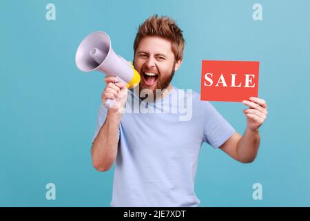 Porträt eines positiv optimistischen, gutaussehenden bärtigen Mannes mit einer Karte mit Verkaufsaufschrift und einem Schreien im Megaphon über Rabatte. Innenaufnahme des Studios isoliert auf blauem Hintergrund. Stockfoto