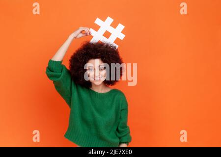 Porträt einer lustigen positiven Frau mit Afro-Frisur, die einen grünen Pullover im lässigen Stil trägt, der Hashtag über ihrem Kopf hält und lächelt. Innenaufnahme des Studios isoliert auf orangefarbenem Hintergrund. Stockfoto