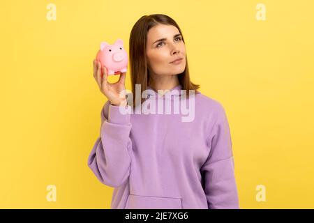 Porträt einer nachdenklichen Frau mit einem angenehmen Aussehen schüttelt Sparschwein, neugierig auf die Höhe ihrer Ersparnisse, trägt lila Hoodie. Innenaufnahme des Studios isoliert auf gelbem Hintergrund. Stockfoto
