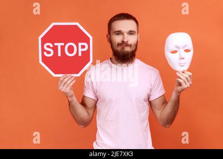 Porträt eines verwirrten, bärtigen Mannes mit weißer Maske, unbekanntem Gesicht und rotem Verkehrsschild, der die Kamera ansah und ein pinkes T-Shirt trug. Innenaufnahme des Studios isoliert auf orangefarbenem Hintergrund. Stockfoto