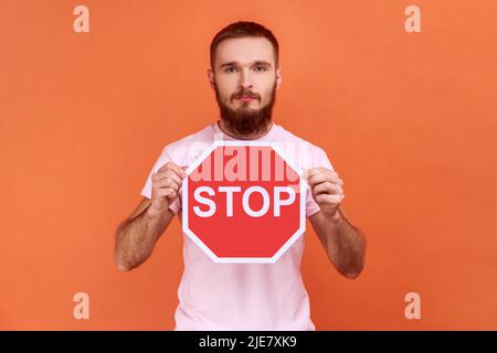 Porträt eines seriös beherrten bärtigen Mannes mit rotem Stoppschild, der mit ernstem Ausdruck die Kamera anschaut und ein pinkfarbenes T-Shirt trägt. Innenaufnahme des Studios isoliert auf orangefarbenem Hintergrund. Stockfoto