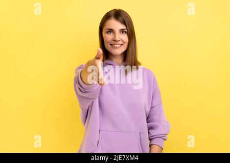 Freut mich, Sie kennenzulernen. Porträt einer freundlich lächelnden Frau, die ihre Hand ausstreckt und Gäste empfängt, Gastfreundschaft und einen violetten Hoodie trägt. Innenaufnahme des Studios isoliert auf gelbem Hintergrund. Stockfoto