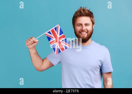 Porträt eines gutaussehenden, zufriedenen bärtigen Mannes, der die Flagge einer konstituierenden Einheit des Vereinigten Königreichs hält und den Tag der britischen Unabhängigkeit feiert. Innenaufnahme des Studios isoliert auf blauem Hintergrund. Stockfoto