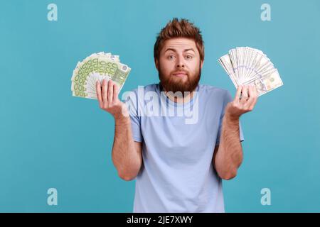 Porträt eines verwirrten, nachdenklichen bärtigen Mannes, der Fans von Dollar- und Euro-Banknoten, Wechselkurse und Zahlungsdiensten hält. Innenaufnahme des Studios isoliert auf blauem Hintergrund. Stockfoto