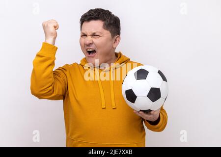 Positiv zufriedener Mann, der den Fußball hält, den Sieg der Lieblings-Fußballmannschaft in der Meisterschaft feiert und einen Hoodie im urbanen Stil trägt. Innenaufnahme des Studios isoliert auf weißem Hintergrund. Stockfoto