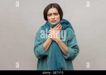 Porträt einer Frau, die in einen warmen Schal gewickelt ist, sich mit Fieber, saisonalen Grippesymptomen und Halsschmerzen unwohl fühlt und eine Jacke im lässigen Stil trägt. Innenaufnahme des Studios isoliert auf grauem Hintergrund. Stockfoto